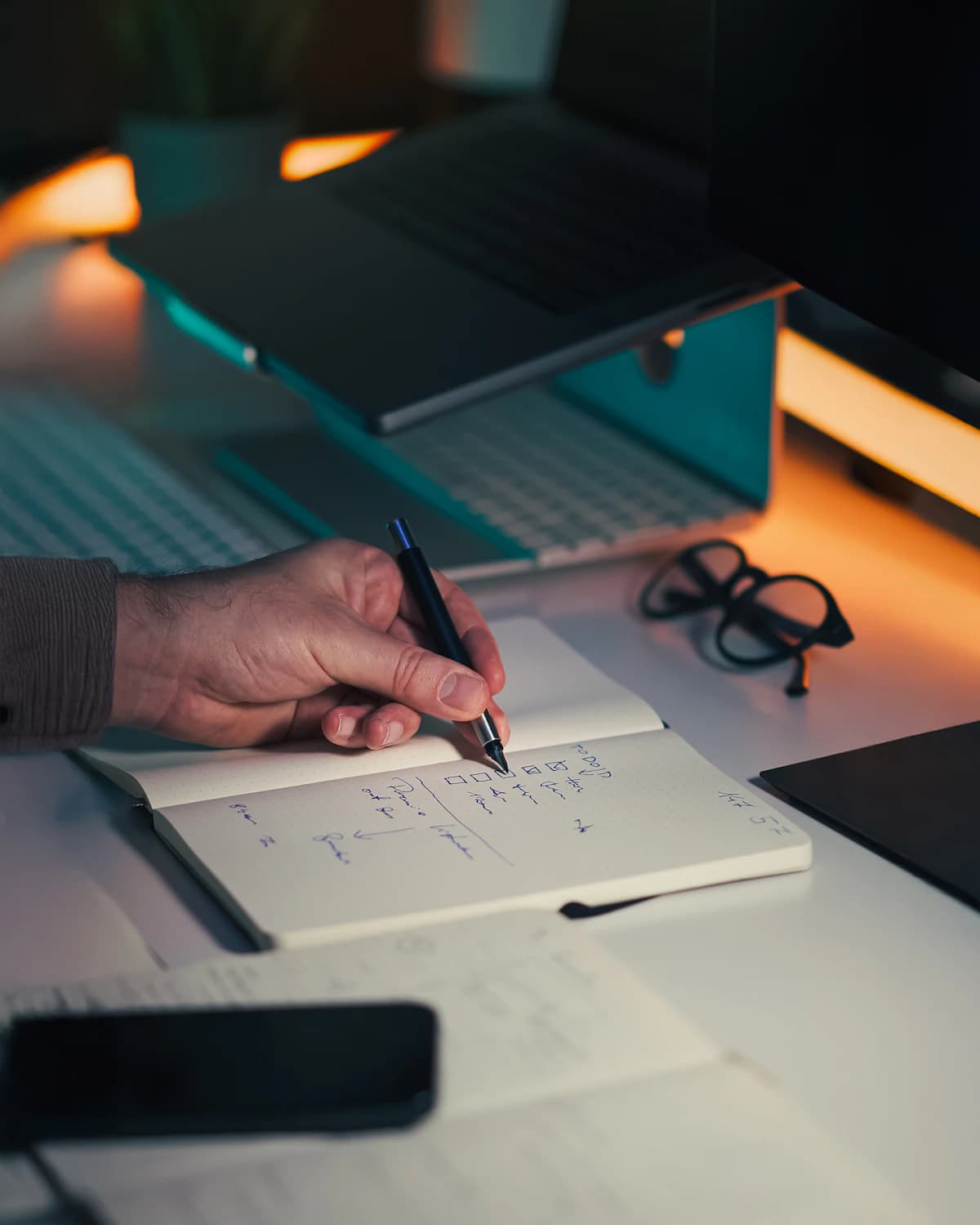 A hand writing notes in a notebook beside a laptop